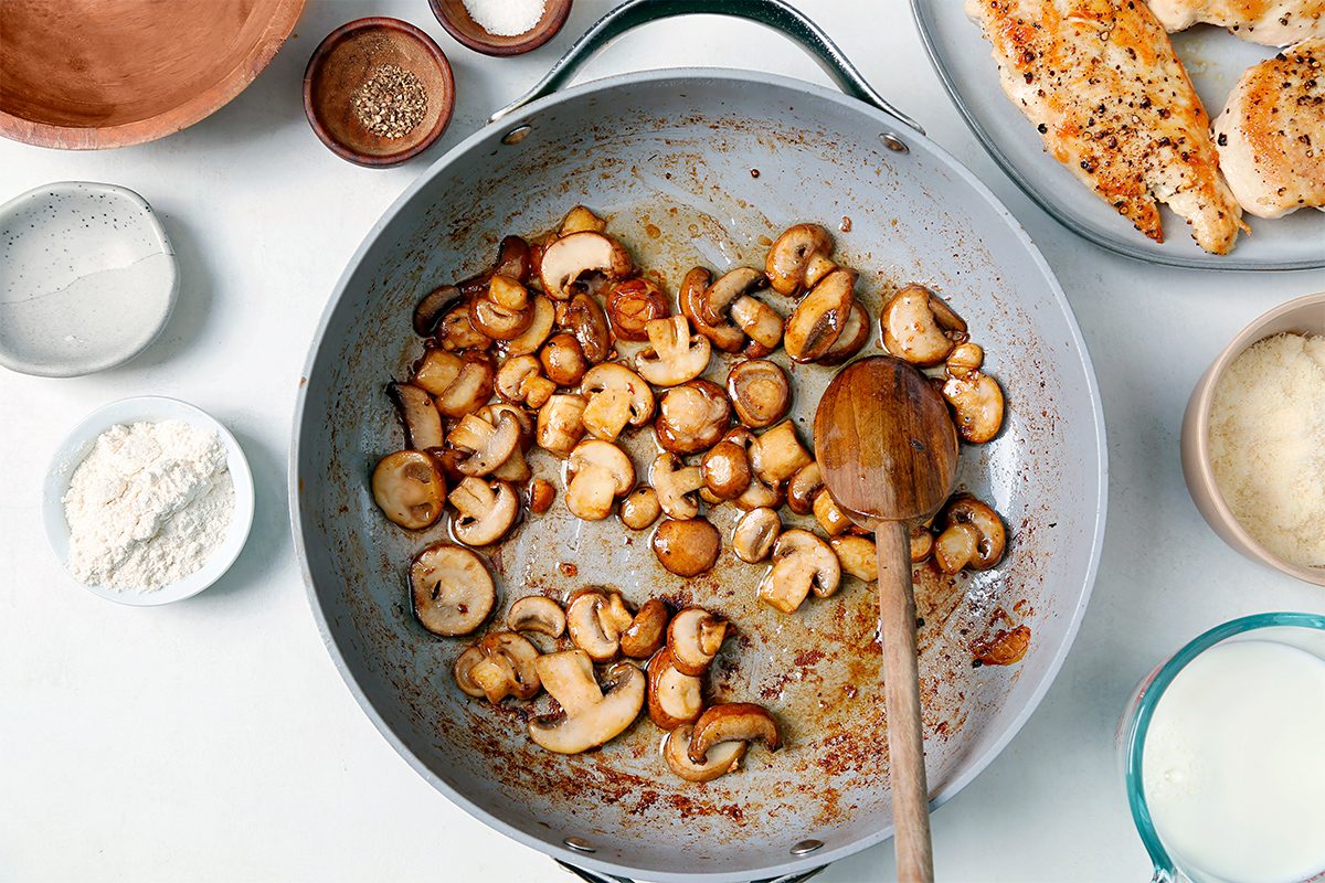 A frying pan contains sautéed mushrooms with a wooden spoon. Surrounding the pan are small bowls of flour, seasoning, and cream, as well as a plate with cooked chicken breasts. The scene is set on a white surface.