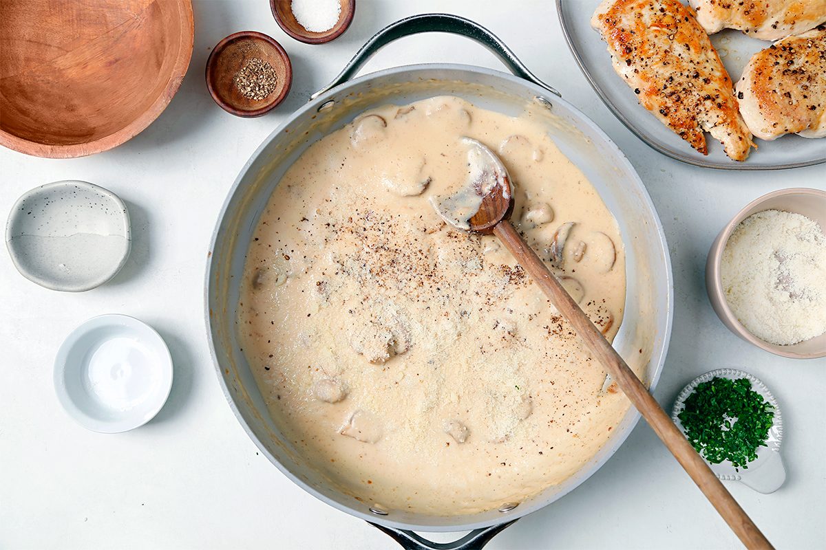 A pan filled with creamy mushroom sauce, featuring visible mushroom pieces, and a wooden spoon. Surrounding the pan are bowls of ingredients, a plate with seasoned chicken, and a sprig of parsley. The setting is a white countertop.