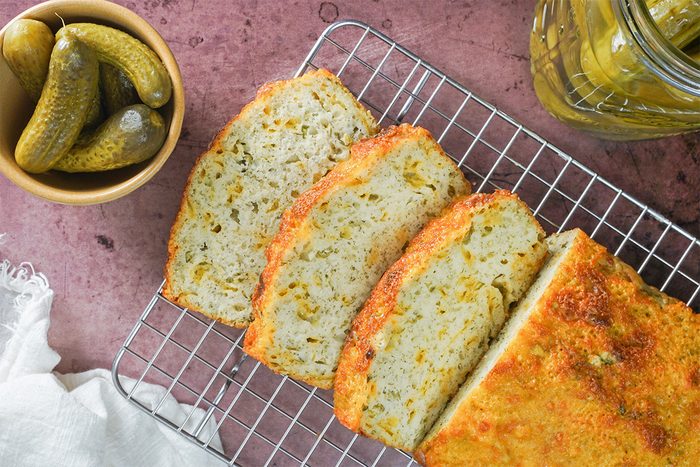 Sliced dill pickle bread on a wire rack with a bowl of pickles and a jar of pickles nearby, all on a pink surface.