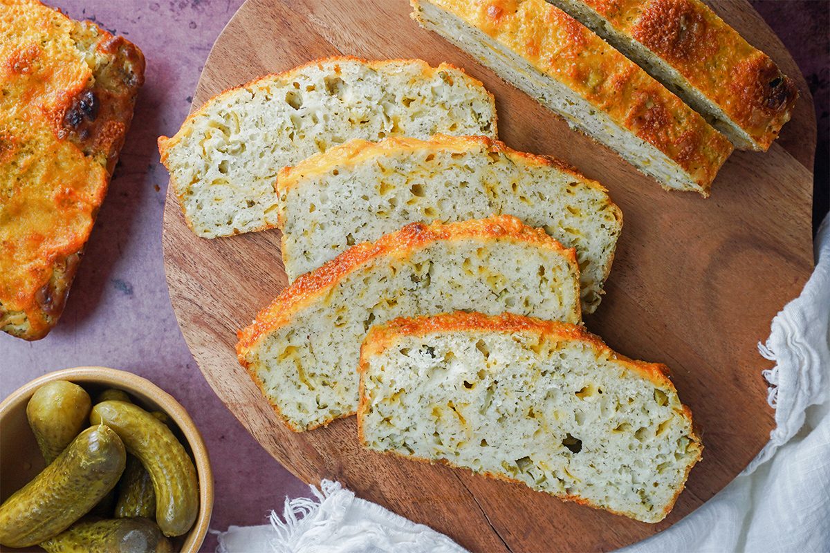 \Sliced loaf of cheese and herb bread on a wooden round board, with pieces arranged neatly. A whole loaf is partially visible, and a small bowl of pickles is placed nearby. A white cloth is draped to the right.