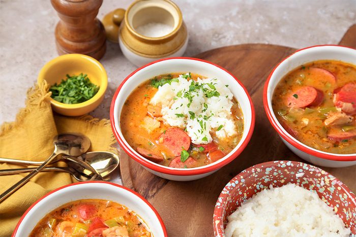 Three bowls of soup with carrots and garnish are placed on a wooden tray. A side bowl of white rice and a small dish of chopped herbs are nearby. Silverware and a yellow napkin are on the side. Salt and pepper shakers complete the setting.