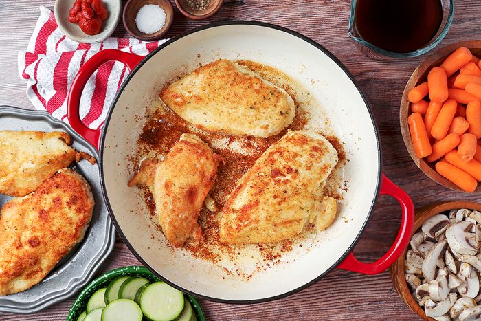 Pan with browned breaded chicken breasts on a wooden table, surrounded by bowls of carrots, mushrooms, zucchini slices, salt, pepper, ketchup, and a jug of sauce. A red-striped towel is partially visible.