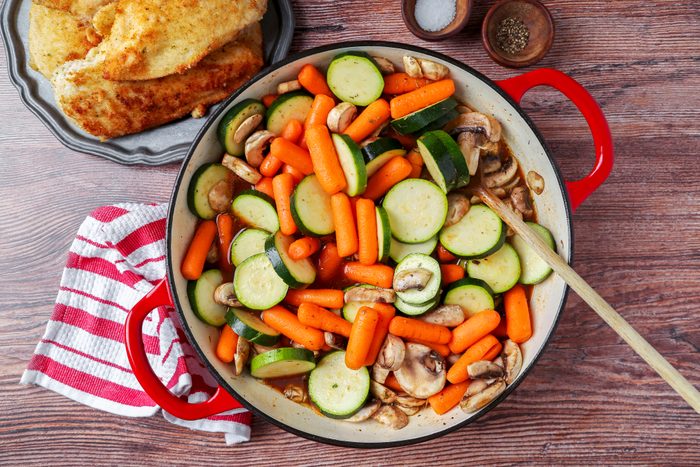 A pan filled with sliced zucchini, carrots, and mushrooms, with a wooden spoon resting inside. A red and white striped cloth lies beside it. In the background, there's a plate of breaded and fried food and small bowls of salt and pepper.