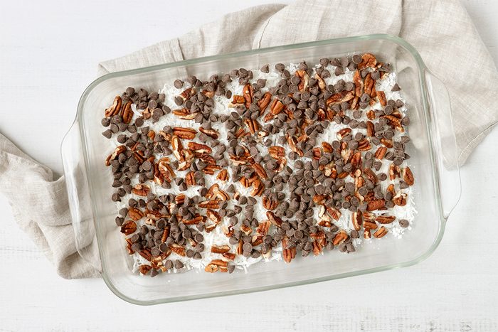 A glass baking dish filled with a dessert topped with chopped pecans and chocolate chips rests on a light-colored surface. A beige cloth is partially tucked under the dish.