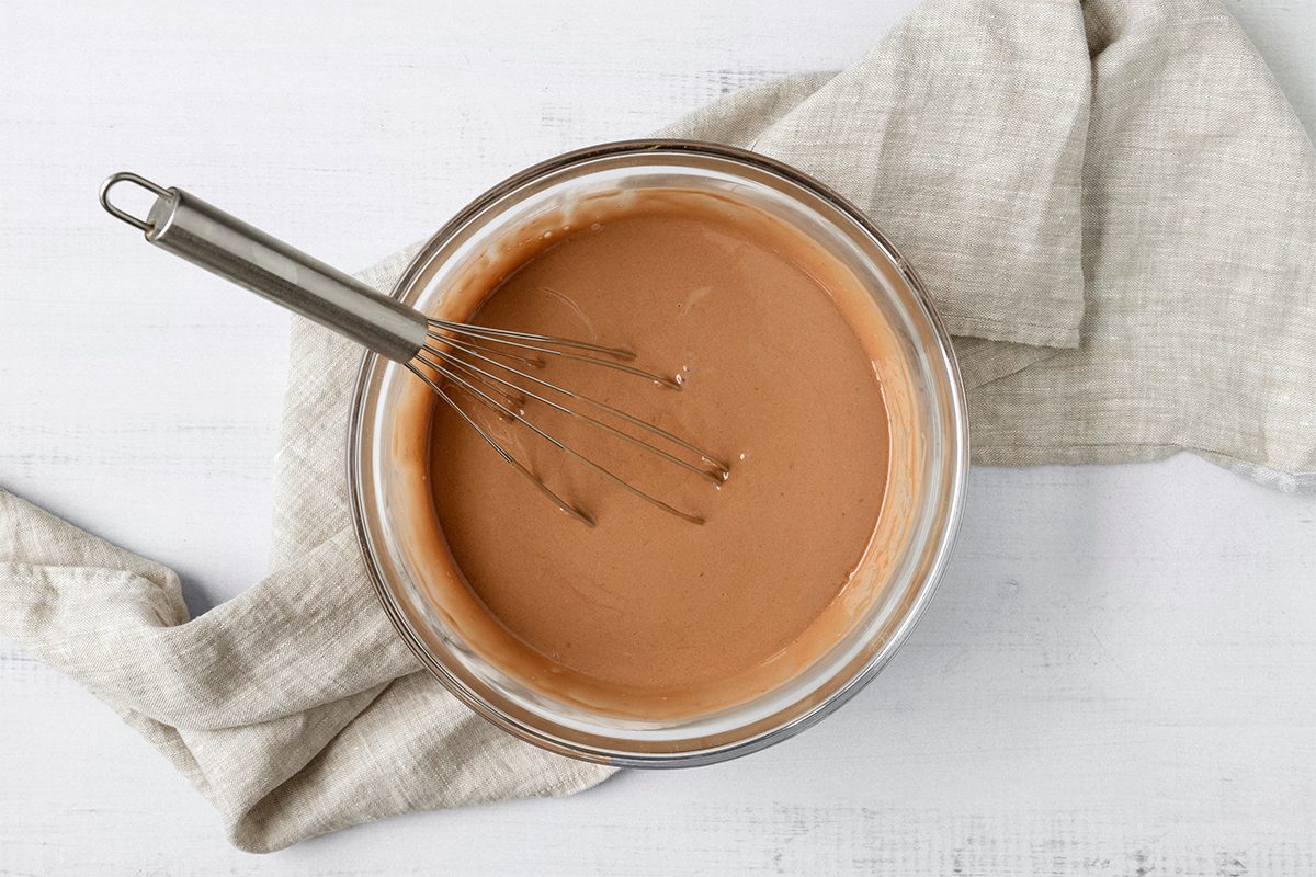 A glass bowl of light brown batter with a whisk in it, resting on a textured, light-colored cloth. The background is a white wooden surface.