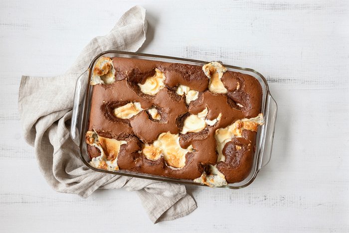 A glass baking dish filled with chocolate brownies topped with swirls of melted marshmallows. A beige cloth napkin is placed beside the dish on a white wooden surface.