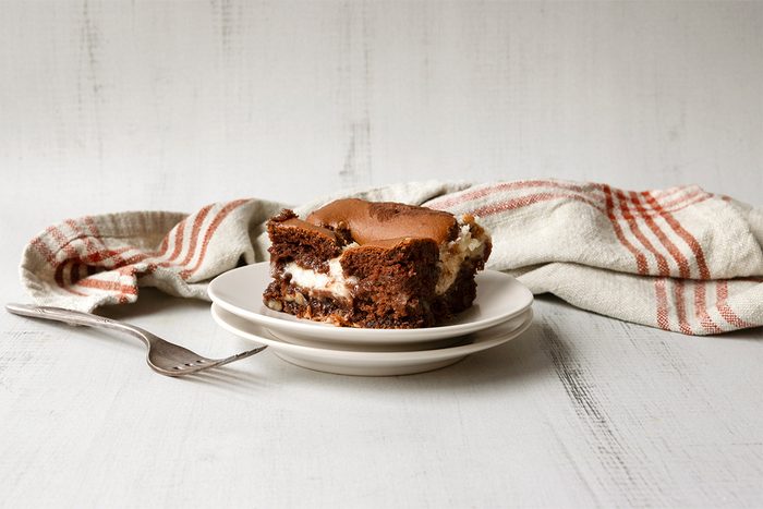 A slice of chocolate cake with cream filling on a white plate. A fork lies beside the plate on a white wooden surface. A striped kitchen towel is in the background.