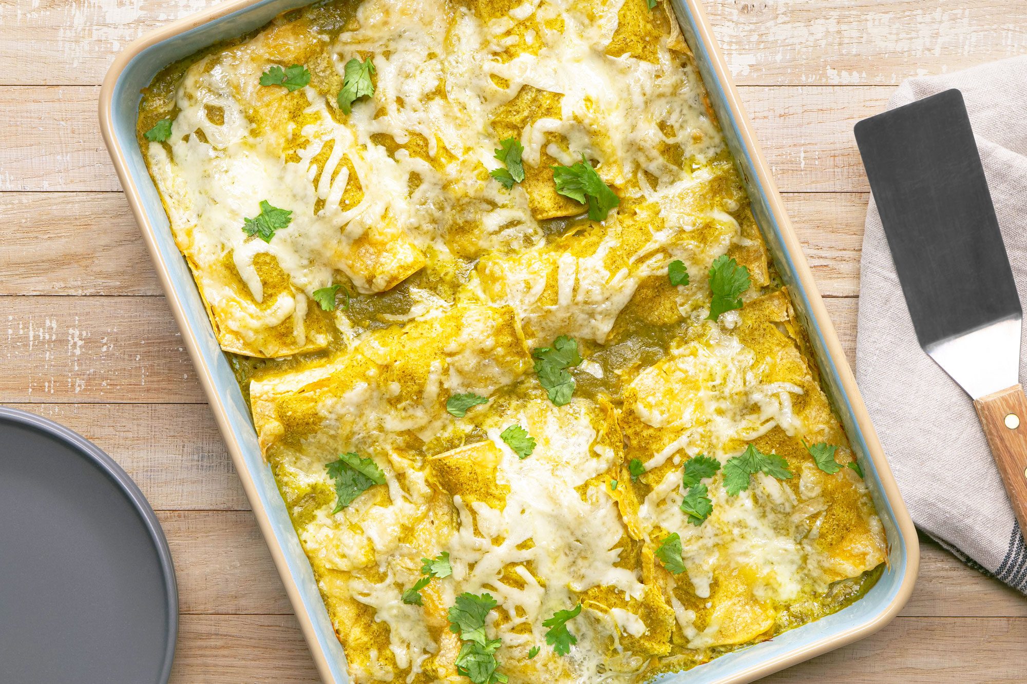 overhead shot of a dish of Enchiladas Verdes; the enchiladas are arranged in a blue and beige baking dish and covered in a creamy green sauce, shredded cheese and fresh cilantro; the dish is on a wooden table with a grey plate, a linen napkin and a spatula
