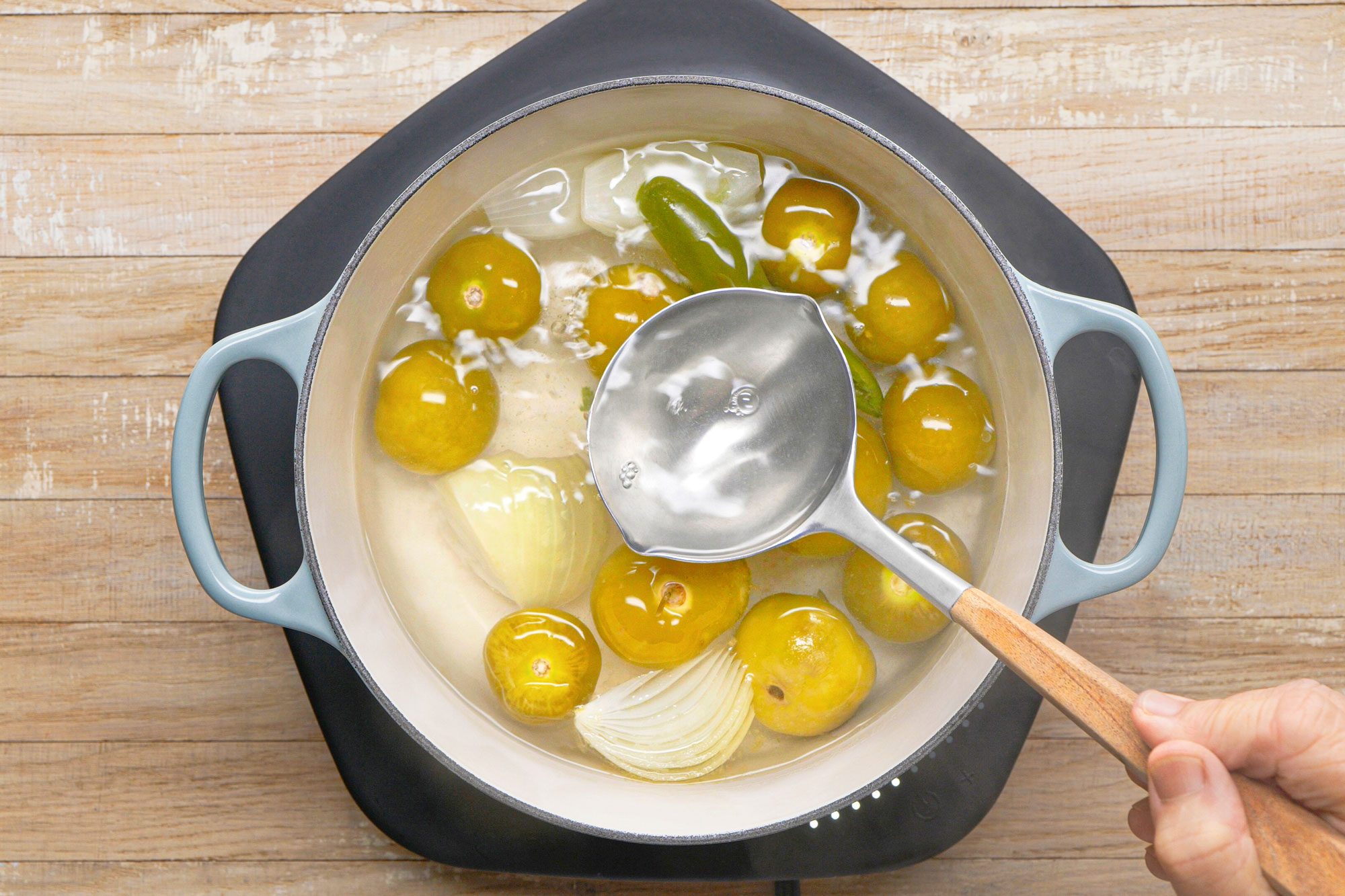 overhead shot of a pot of simmering liquid on a stovetop burner, with a ladle being used to stir the contents, The pot is light blue with black accents and has a wooden handle; The ladle is also wooden, with a metal bowl; The pot contains a mixture of yellow and green vegetables, including tomatillos and onions