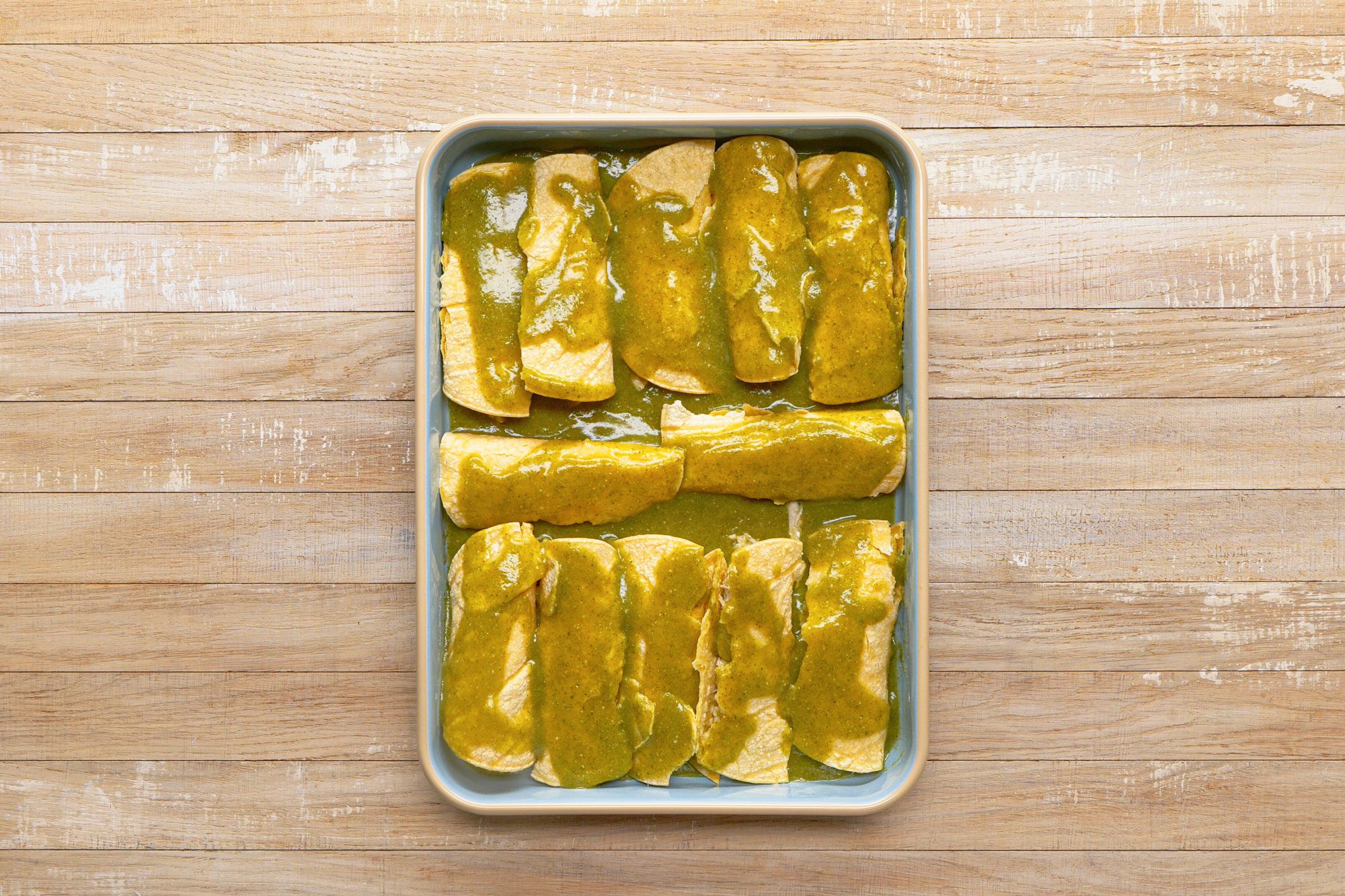 overhead shot of a baking dish containing enchiladas; the enchiladas are covered in a green sauce,the baking dish is on a wood surface