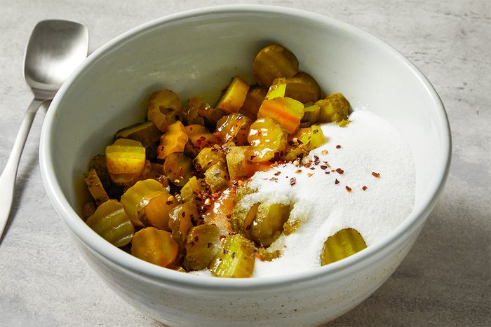 A bowl filled with sliced pickles, a white powder, and sprinkled spices, accompanied by a spoon on the side.