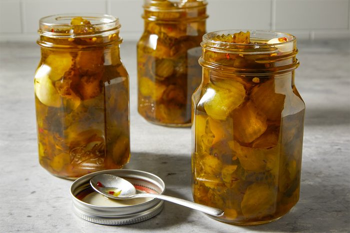 Three glass jars filled with preserved vegetables, likely pickles, are placed on a gray countertop. A lid and a spoon rest nearby. The jars are filled with chunks of vegetables in a yellowish-brown liquid.