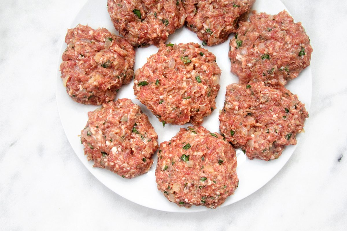 Eight raw hamburger patties with visible herbs and seasonings are arranged on a white plate set against a marble background.