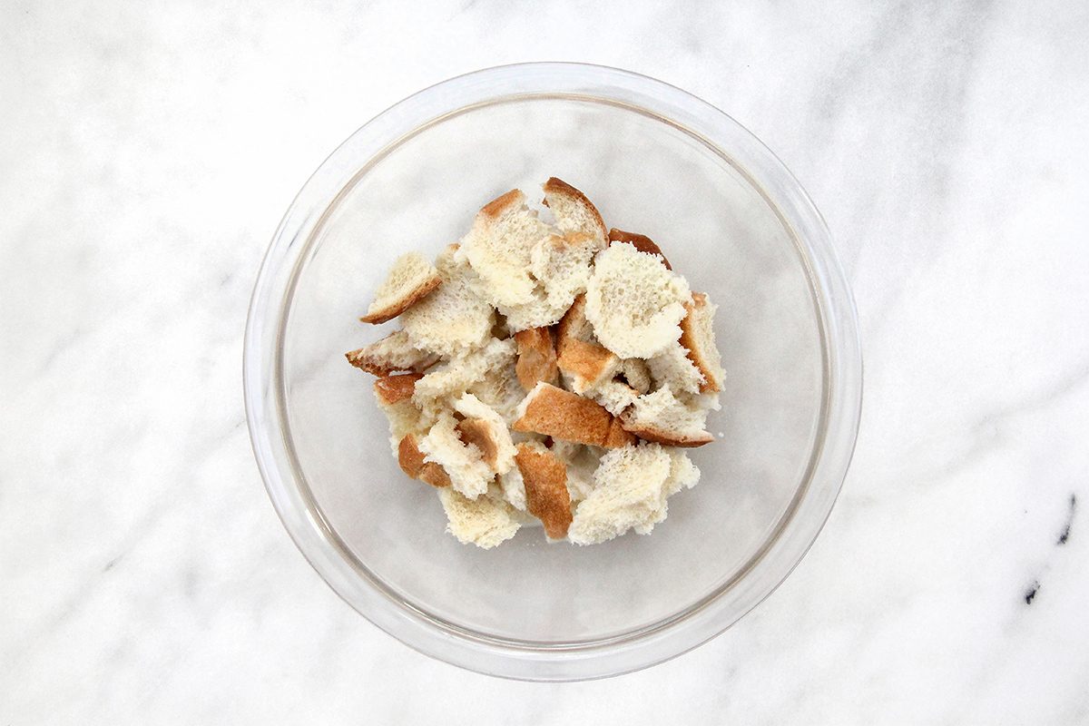 A clear glass bowl filled with torn pieces of bread sits on a white marble surface. The bread pieces are a mix of crust and soft interior.