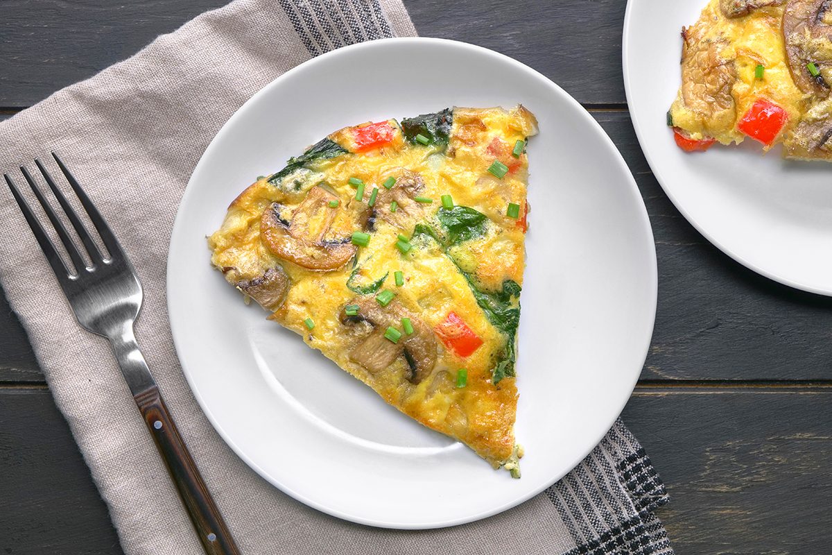 A slice of vegetable frittata on a white plate, featuring mushrooms, spinach, and red bell peppers. The frittata is garnished with chopped green herbs. A fork rests on a folded cloth napkin beside the plate, set on a dark wooden surface.
