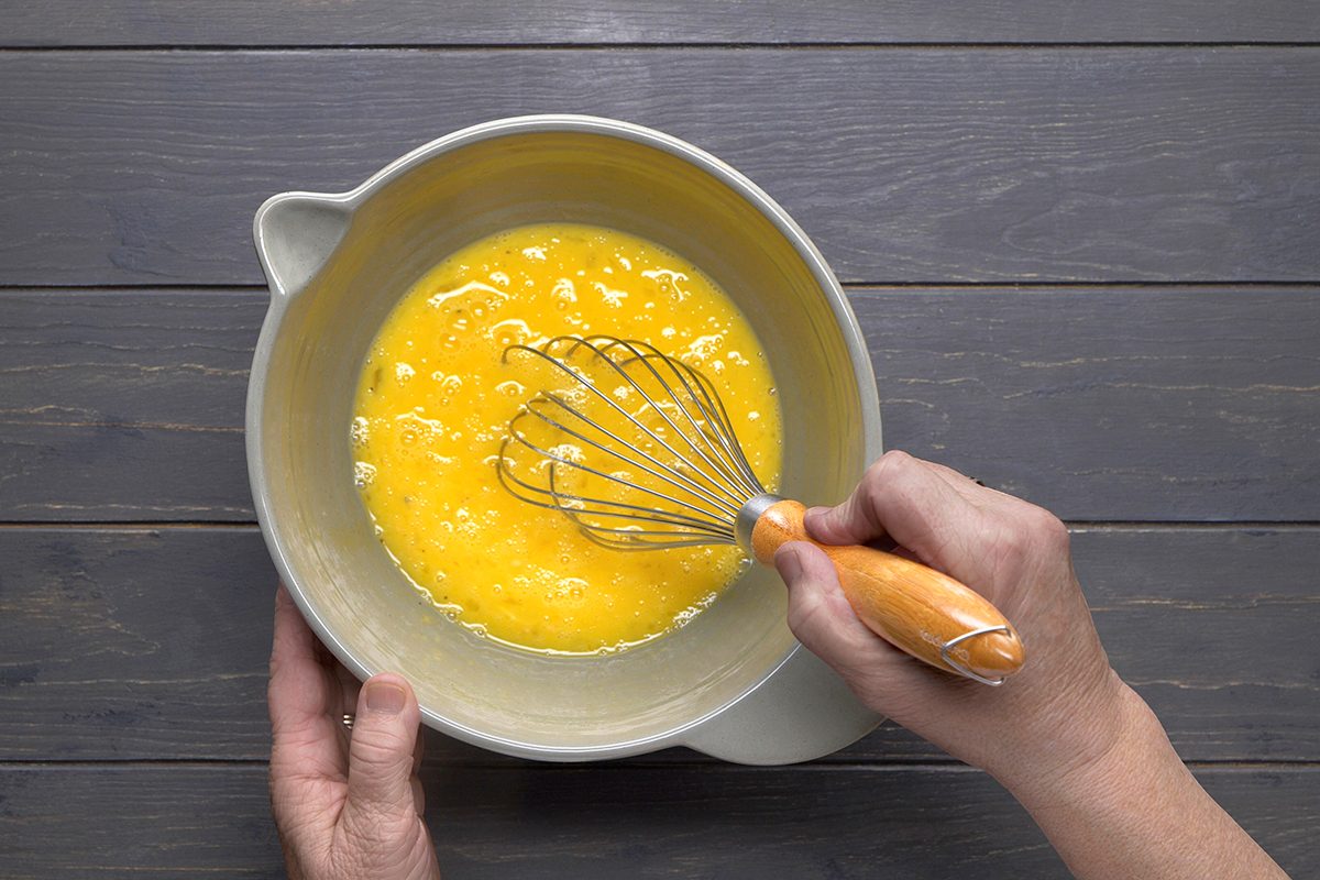 Hands whisking eggs in a gray mixing bowl on a wooden surface.