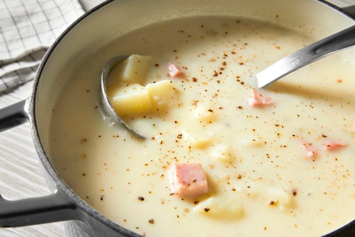 closeup shot of a pot of Ham and Potato Soup; a silver ladle is resting on the edge of the pot; the pot is sitting on a white table and a white and gray checkered cloth is visible to the left of the pot