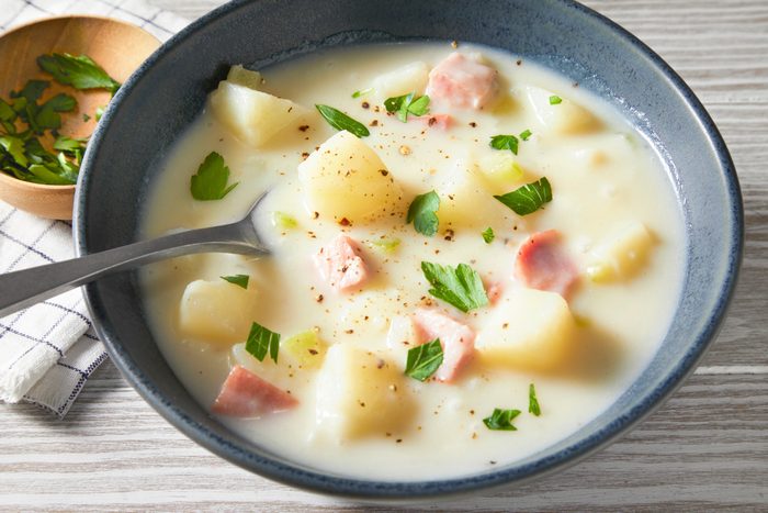 3/4th shot of A bowl of Ham and Potato Soup, a few sprigs of parsley are scattered throughout; the bowl is sitting on a light wood table, there is a small bowl of chopped parsley and a cloth napkin to the left of the soup bowl