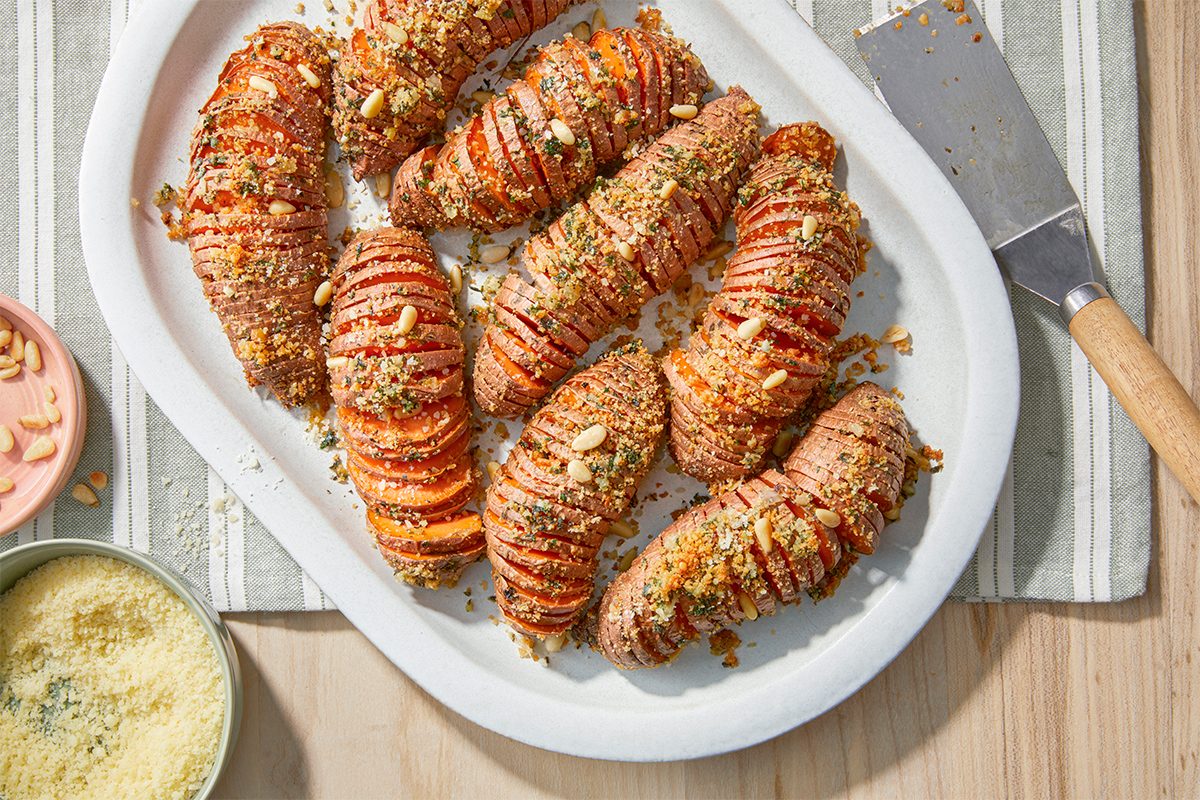A white platter holds several roasted Hasselback sweet potatoes topped with a breadcrumb and herb mixture. A small bowl of grated cheese and a knife are nearby on a striped cloth.