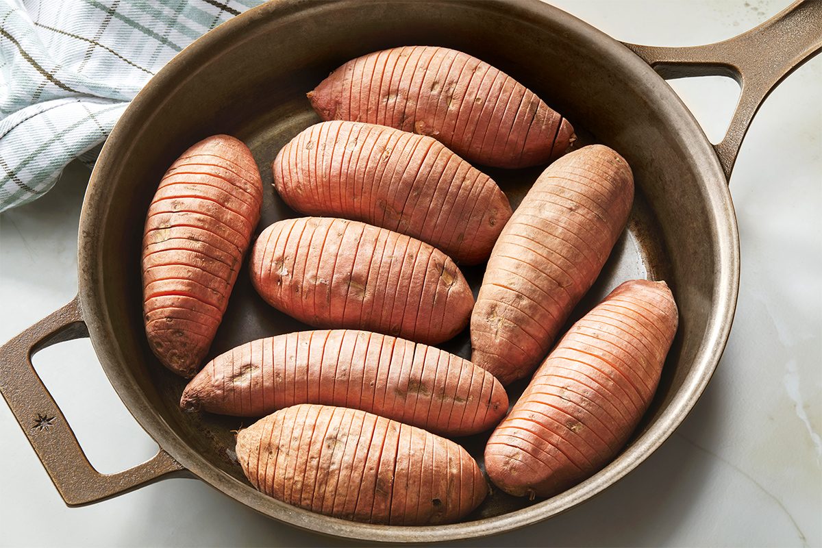 A round baking dish holds nine uncooked, scored sweet potatoes. A checkered kitchen towel is partially visible in the background on the left side.