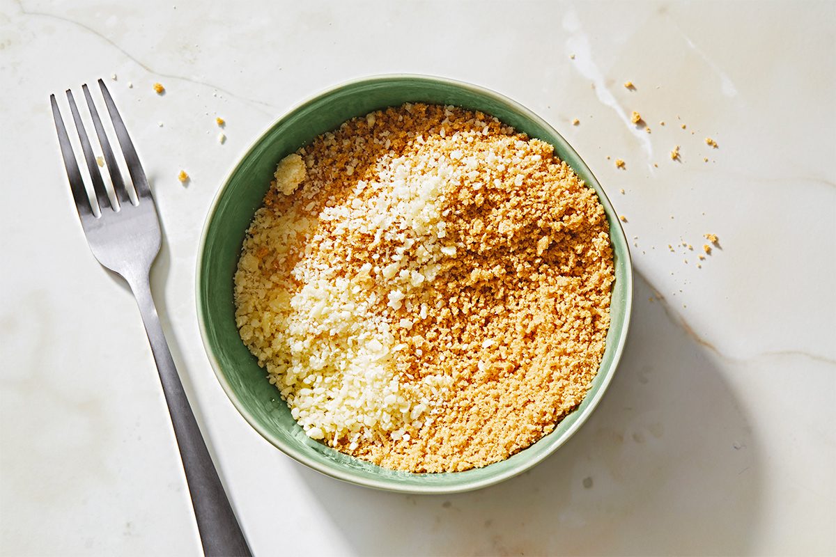 A bowl filled with grated Parmesan and cheddar cheese sits on a marble surface. A fork is placed to the left of the bowl. The cheese contrasts in color, with yellow cheddar and pale Parmesan creating a distinct division within the bowl.