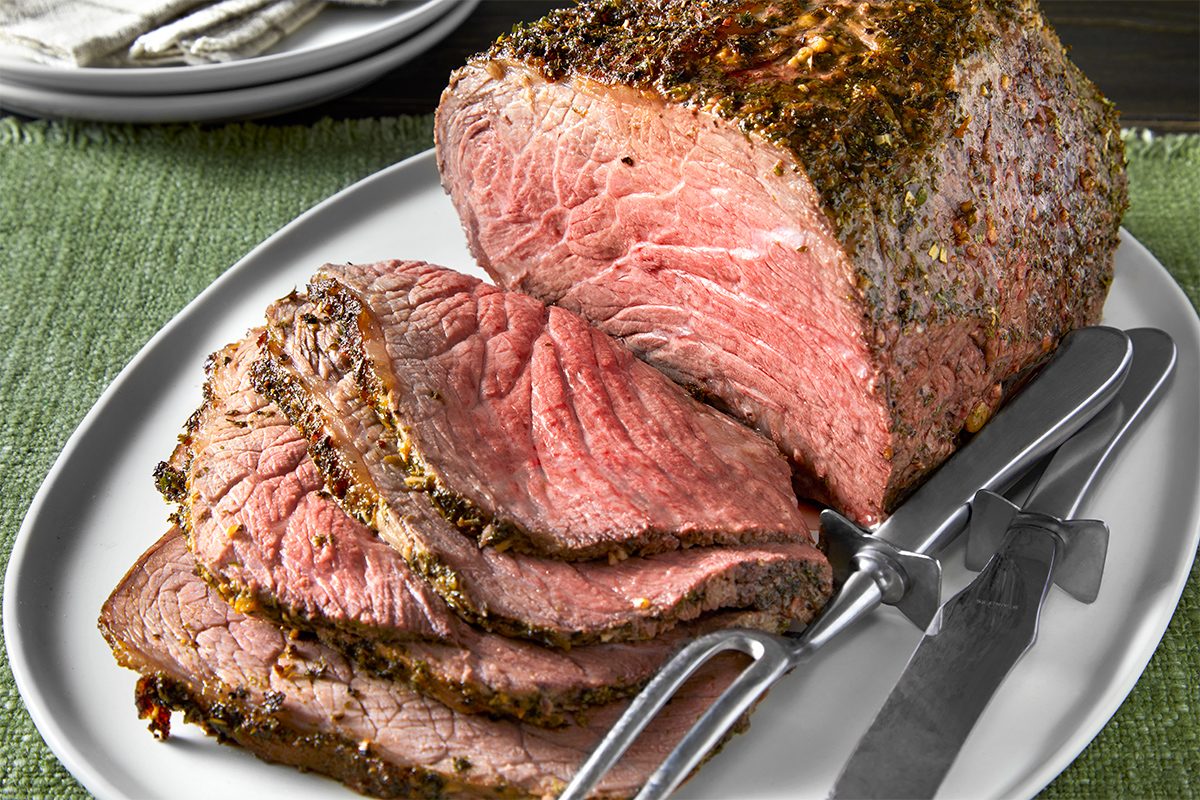 Close shot of Herb-Crusted Roast Beef; served on large plate; knife and fork; napkin; serve with gravy; table cloth; dark wooden surface;
