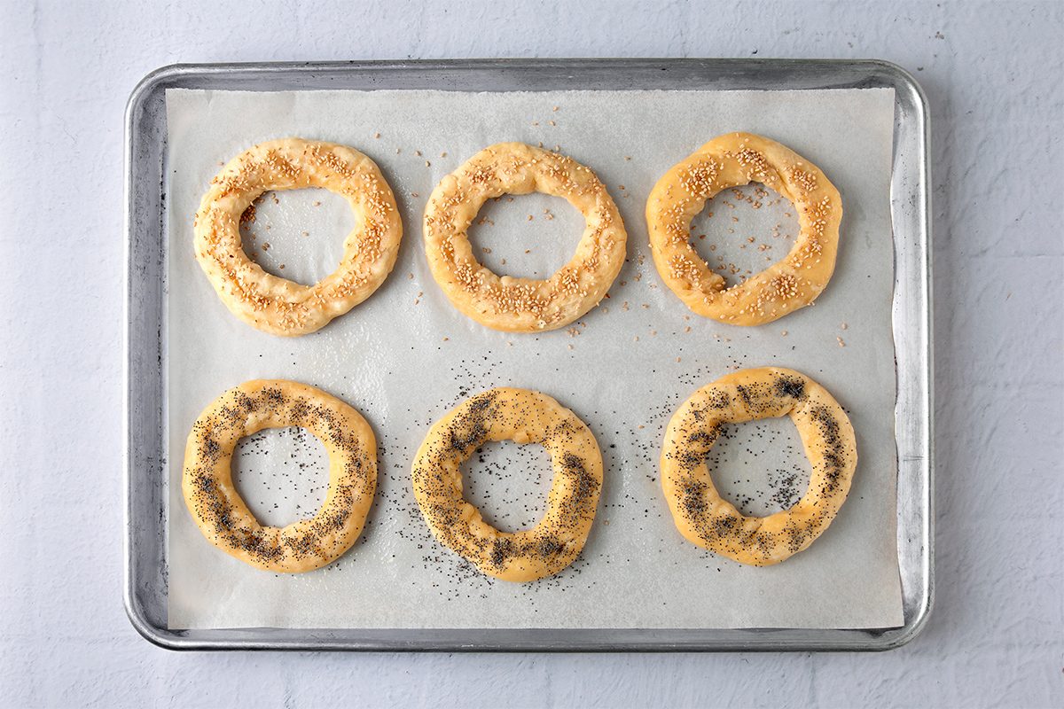 Six raw circular dough rings sprinkled with seeds are arranged neatly on a parchment-lined baking sheet, ready for baking. The baking sheet rests on a light gray surface.