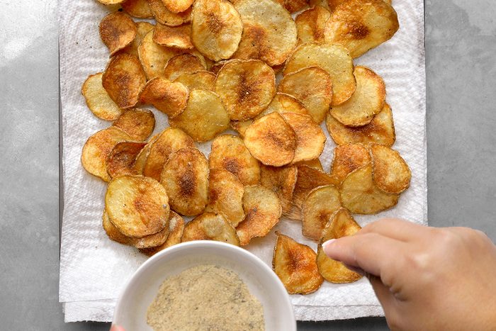 A person sprinkles seasoning from a bowl onto homemade potato chips spread on a paper towel. The chips are golden brown and freshly cooked, placed on a light gray surface.