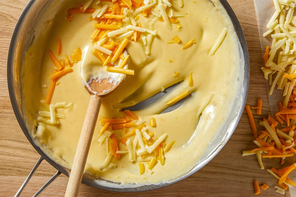 A pan of creamy cheese sauce with shredded orange and white cheese partially mixed in. A wooden spoon rests inside the sauce, with more shredded cheese scattered on the wooden surface nearby.
