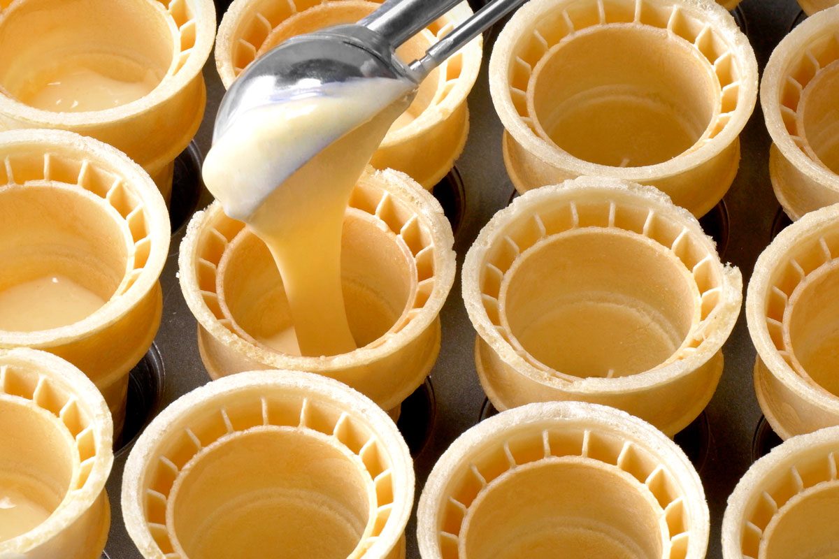 a close-up view of an array of waffle cones; the cones are arranged in rows; a metal ice cream scoop filled with a light yellow substance is suspended above the cones, with the liquid being poured into one of the cones;