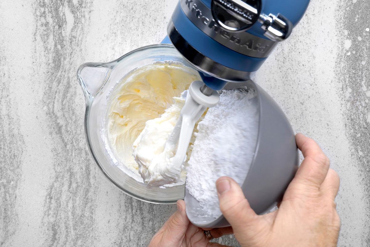 a person adding powdered sugar to a bowl of butter cream frosting in a stand mixer; a blue stand mixer is attached to a glass mixing bowl containing whipped butter cream frosting