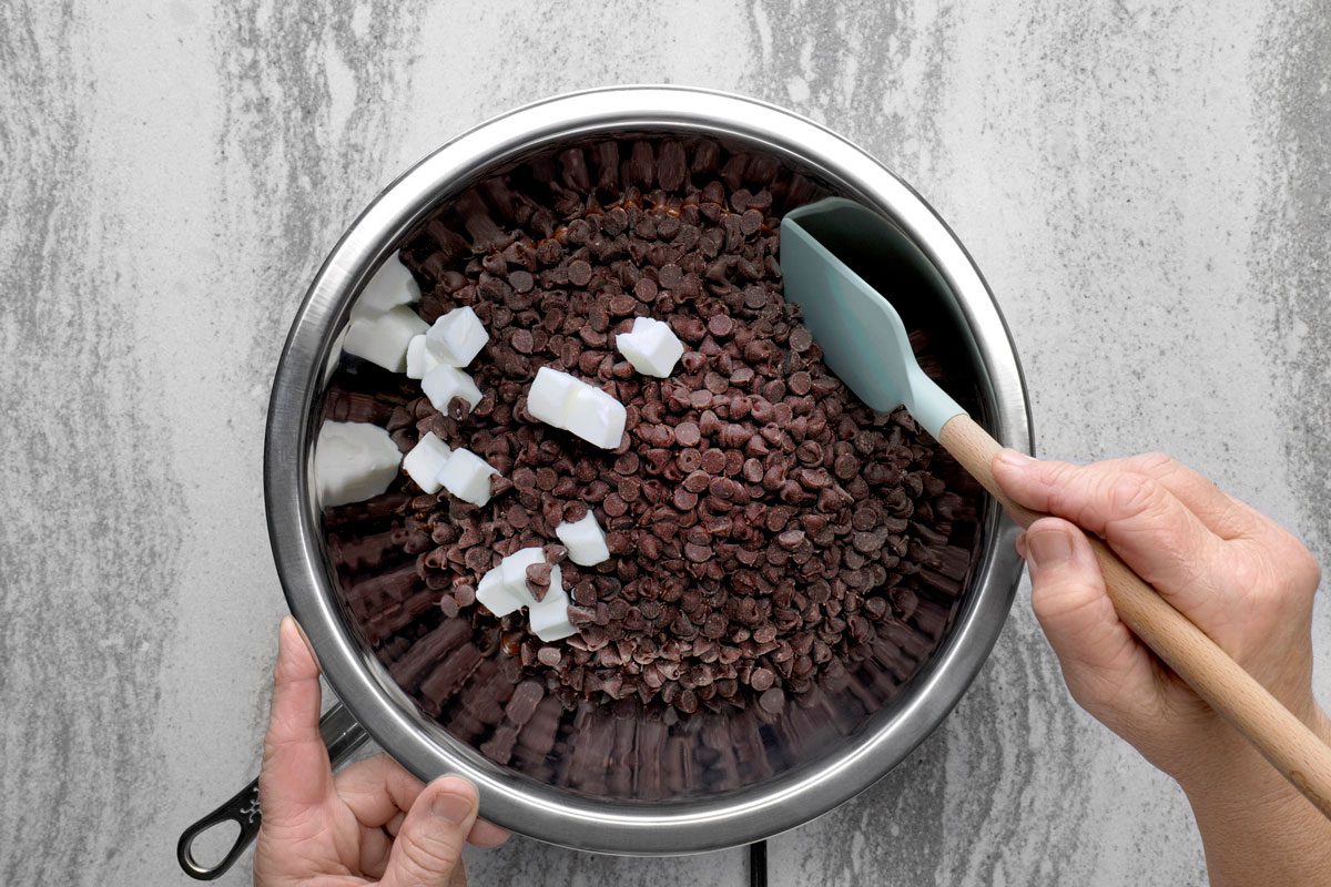 overhead shot of a stainless steel bowl filled with chocolate chips and white chocolate chunks; a blue spatula with a wooden handle is placed inside the bowl; the bowl is being held by a person's hand; the background is a light gray countertop