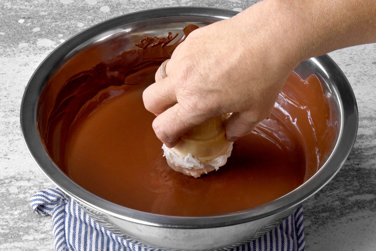 3/4th shot of a hand is dipping an ice cream cone into a bowl of melted chocolate; the hand is holding the cone by the top and dipping the bottom into the chocolate; the bowl is sitting on a white and blue striped kitchen towel, on a speckled grey countertop