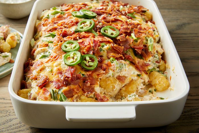 A white casserole dish filled with a baked cheesy tater tot casserole. The top is garnished with crispy bacon pieces and sliced green jalapeños. The dish is placed on a wooden table.