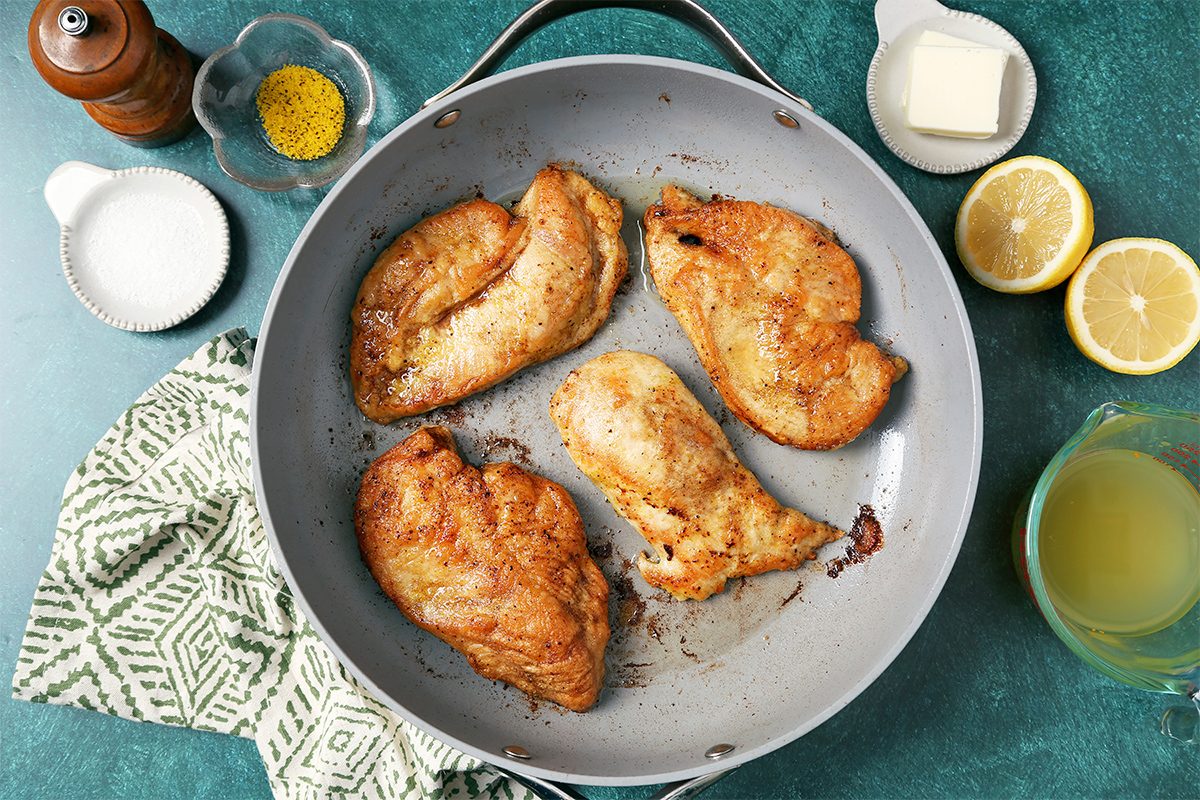 Four cooked chicken breasts in a frying pan on a green surface, surrounded by ingredients like lemon halves, butter, seasoning, and a small bowl of broth. A patterned cloth is nearby.