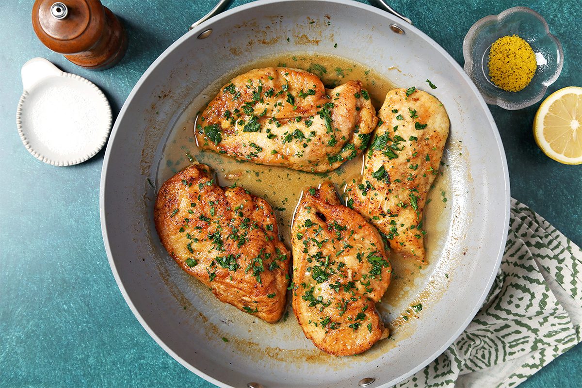 A pan with four golden-brown garlic butter chicken breasts garnished with parsley. A lemon half, a small dish with spices, and a salt shaker are nearby on a green countertop.
