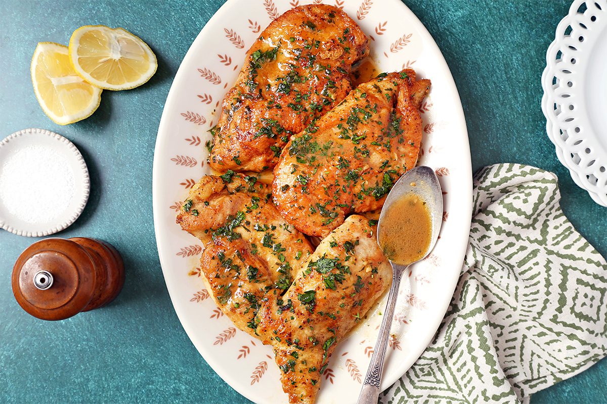 Plate of cooked chicken breasts garnished with herbs and served with a spoon of sauce. Slices of lemon and a small bowl of seasoning are on the side. The dish is on a green surface with a patterned napkin nearby.