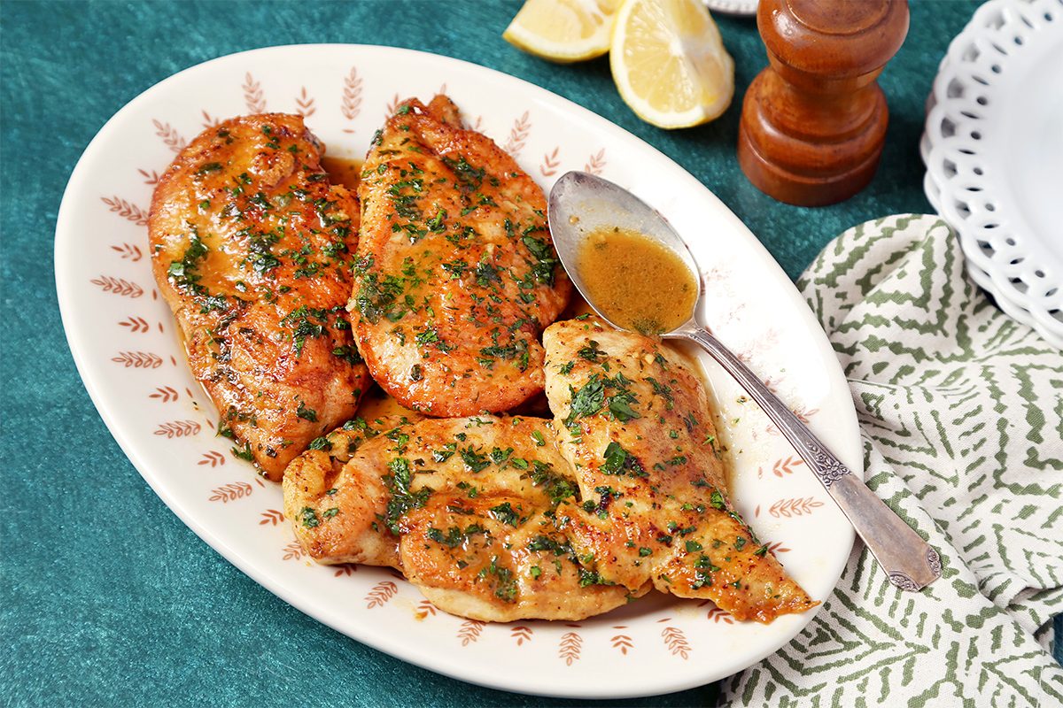 Grilled chicken breasts garnished with herbs are served on an oval plate with a spoon. Lemon wedges and a pepper grinder are in the background on a green tablecloth. A white patterned napkin and plate are nearby.