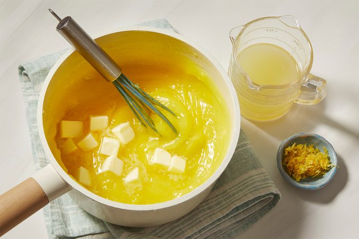 A saucepan with lemon curd being whisked, butter cubes melting. Beside it, a measuring jug with lemon juice and a small bowl of lemon zest. A striped towel is underneath.