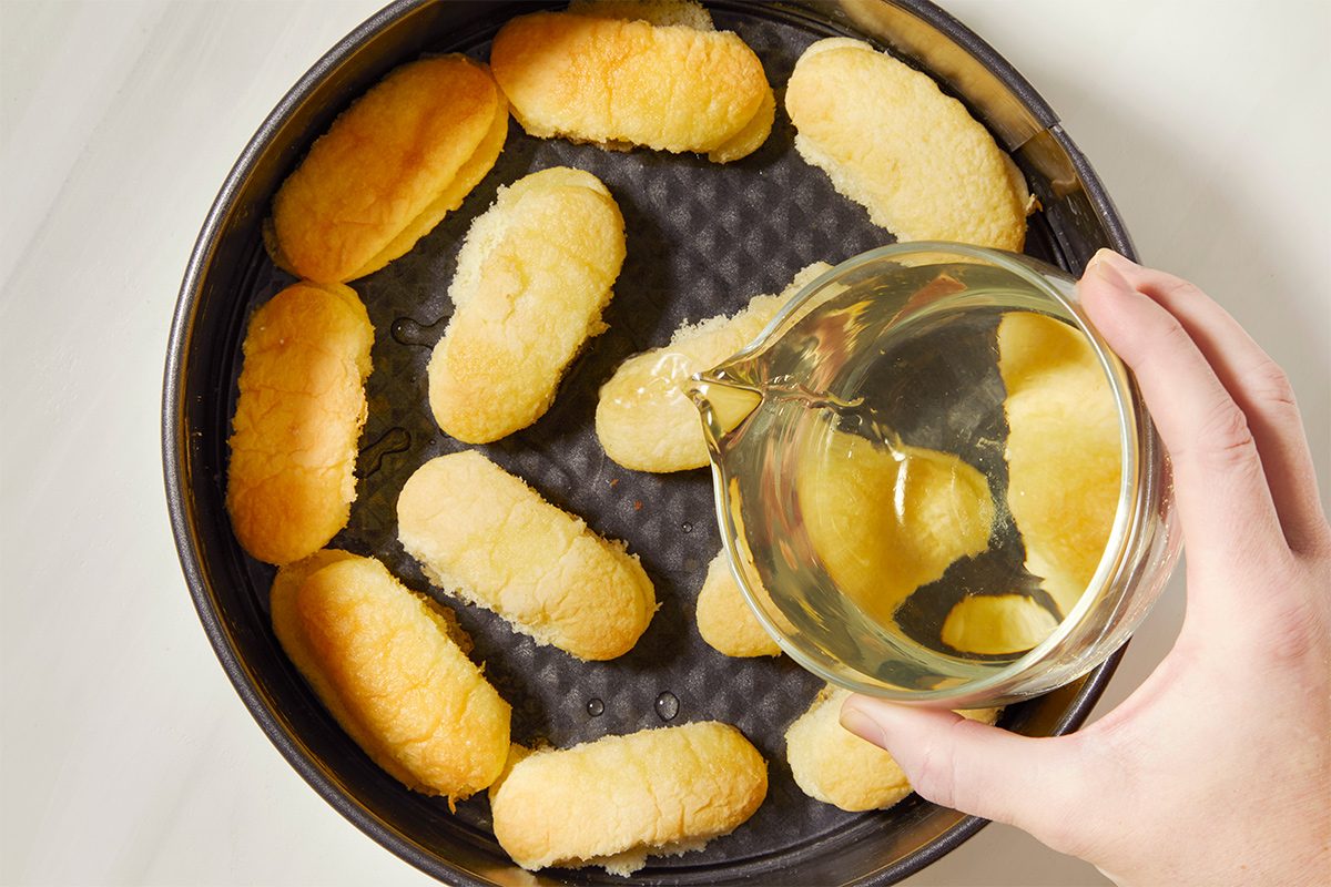 A hand pours a limoncello over ladyfinger cookies arranged in a round baking tin.