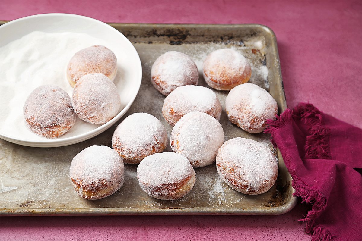 A tray of sugar-coated doughnuts, some placed on a white plate with sugar. The tray and plate are on a pink surface, accompanied by a crumpled burgundy cloth.