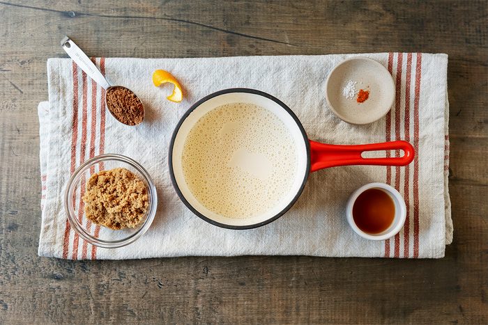 A red-handled saucepan with creamy liquid, surrounded by brown sugar in a glass bowl, vanilla in a small bowl, a dish with a pinch of spice, and a spoon of brown sugar. All items are on a striped cloth on a wooden surface.
