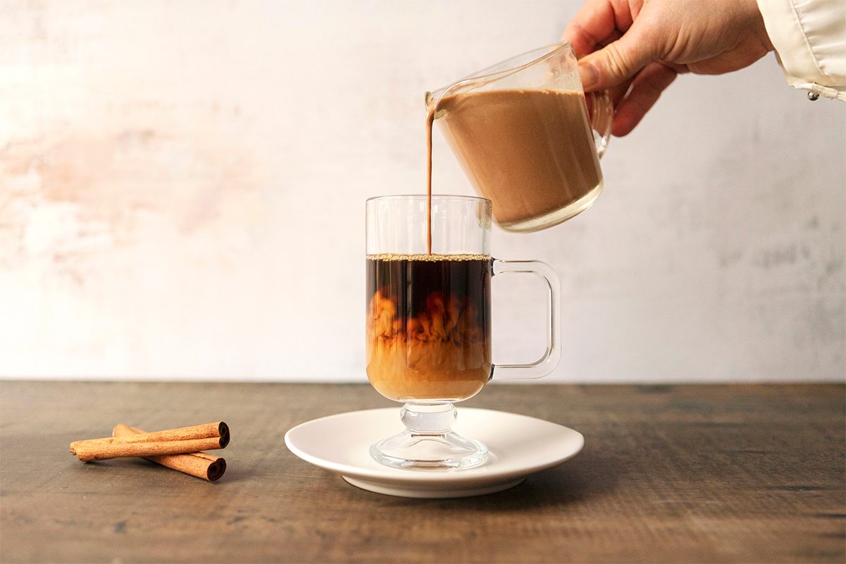 A hand pours milk into a glass cup of coffee on a saucer. Three cinnamon sticks are on the wooden table beside it. The background is a soft white and beige.