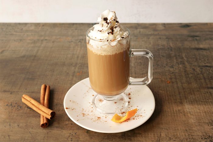 A glass mug of coffee topped with whipped cream and chocolate shavings sits on a white plate with a wooden table. Two cinnamon sticks and a small orange peel are placed on the plate beside the mug.
