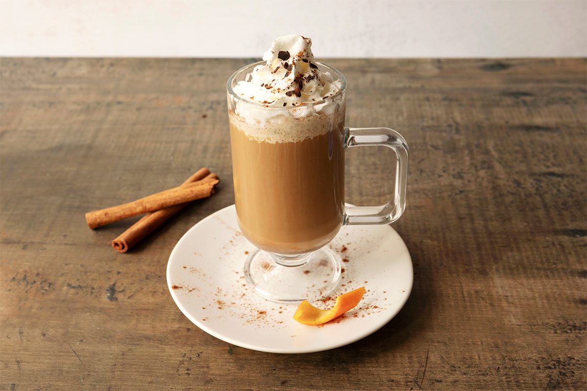 A frothy latte topped with whipped cream and chocolate sprinkles in a clear glass mug on a saucer. Cinnamon sticks and an orange peel garnish the plate on a rustic wooden table.