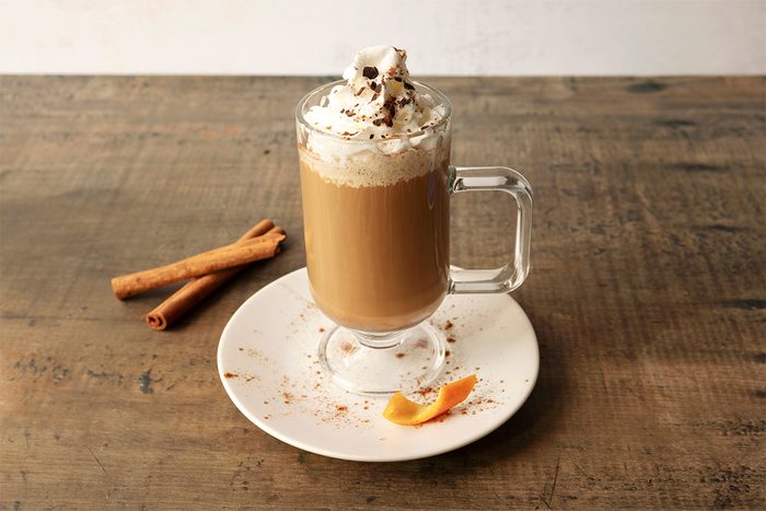 A frothy latte topped with whipped cream and chocolate sprinkles in a clear glass mug on a saucer. Cinnamon sticks and an orange peel garnish the plate on a rustic wooden table.
