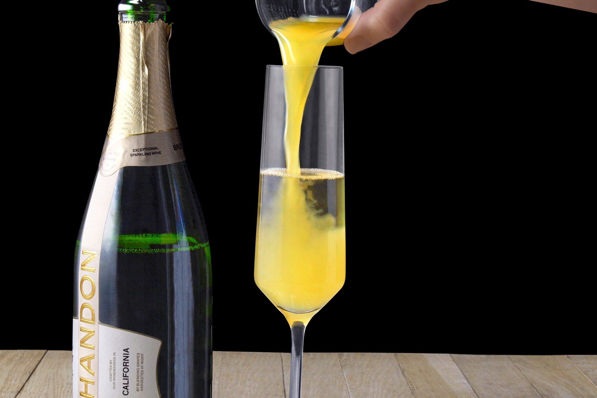 horizontal shot of a person pouring a yellow liquid from a glass pitcher into a glass; a bottle of sparkling wine is in the background on a wooden surface;