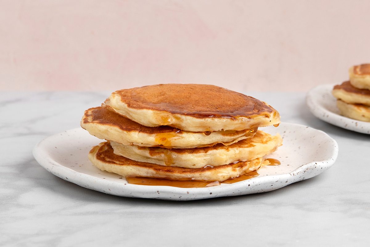 A stack of four golden-brown pancakes with syrup drizzled over them, served on a white speckled plate. The background is a light, blurred surface, creating a cozy breakfast setting.