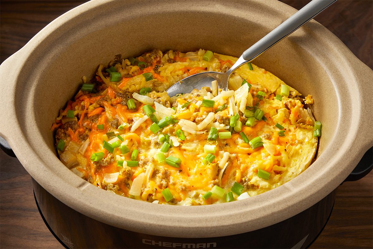 A slow cooker filled with a colorful vegetable and egg dish, garnished with diced green onions. A spoon is resting inside, ready to serve. The dish appears vibrant and freshly cooked.