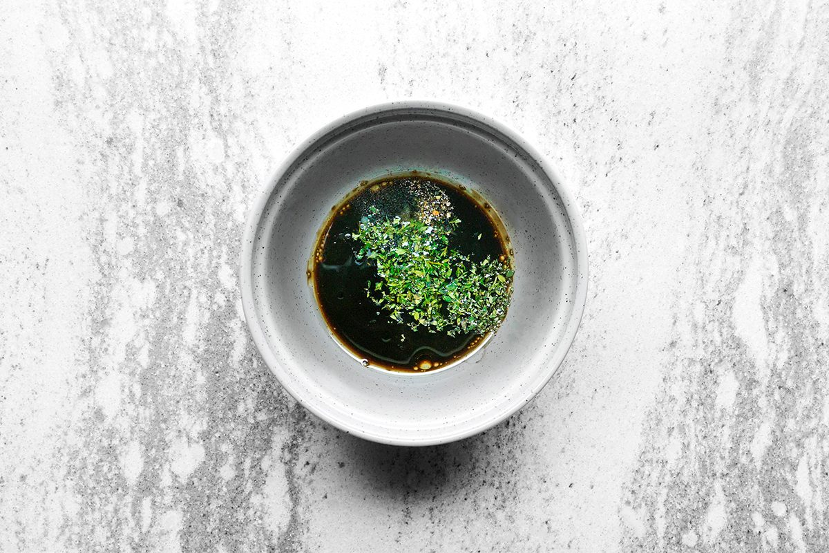A small white bowl filled with dark soy sauce garnished with chopped green herbs, placed on a gray marbled surface.